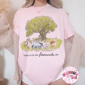 K&ouml;nnte beinhalten: Hellrosa T-Shirt mit einer Cartoon-Illustration von Winnie Puuh und seinen Freunden unter einem Baum. Der Text "Today is my new favourite day" ist in Schreibschrift geschrieben. Das T-Shirt hat einen Rundhalsausschnitt.