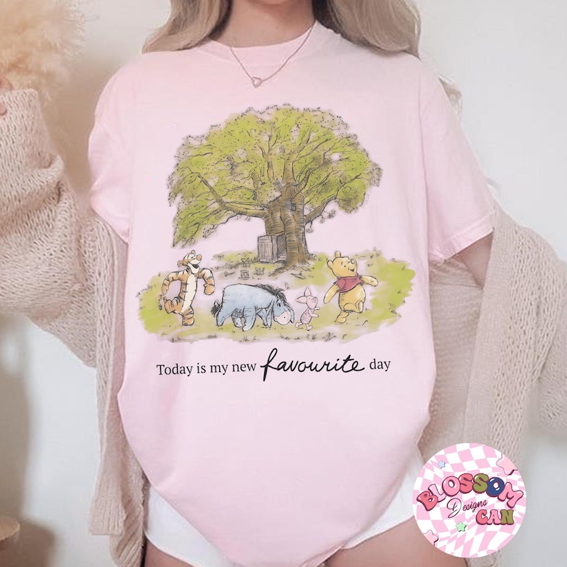 K&ouml;nnte beinhalten: Hellrosa T-Shirt mit einer Cartoon-Illustration von Winnie Puuh und seinen Freunden unter einem Baum. Der Text "Today is my new favourite day" ist in Schreibschrift geschrieben. Das T-Shirt hat einen Rundhalsausschnitt.