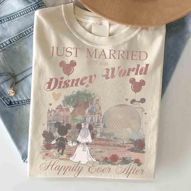 May include: Cream-colored t-shirt featuring the text "Just Married Disney World" with Mickey and Minnie Mouse illustrations. The design includes a castle, Spaceship Earth, and the phrase "Happily Ever After."