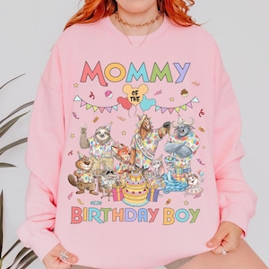 K&ouml;nnte beinhalten: Rosa Sweatshirt mit dem Text "Mommy of the Birthday Boy" und einer farbenfrohen Grafik von Zeichentieren um eine Geburtstagstorte. Das Design beinhaltet Luftballons, Konfetti und Sterne. Das Sweatshirt ist ein Rundhals-Stil.