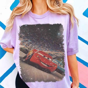Puede incluir: Camiseta lila claro con un estampado del personaje de dibujos animados Rayo McQueen de la pel&iacute;cula Cars. La imagen muestra a Rayo McQueen compitiendo en una pista con otros coches y una multitud de fondo.