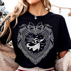 May include: Black t-shirt featuring a white graphic from "Tim Burton's The Nightmare Before Christmas." The design includes a heart-shaped frame with a bow, Zero the ghost dog, bats, and the film's title. The shirt is a crew neck.