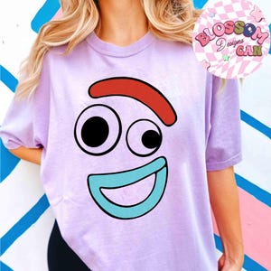 May include: A light purple t-shirt featuring a cartoon face with a red eyebrow, two black eyes, and a blue mouth. The shirt has short sleeves and the text "Blossom Designs Can" in a circular design.