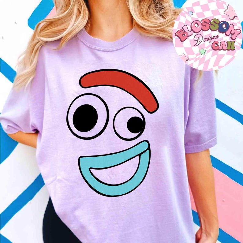 May include: A light purple t-shirt featuring a cartoon face with a red eyebrow, two black eyes, and a blue mouth. The shirt has short sleeves and the text "Blossom Designs Can" in a circular design.