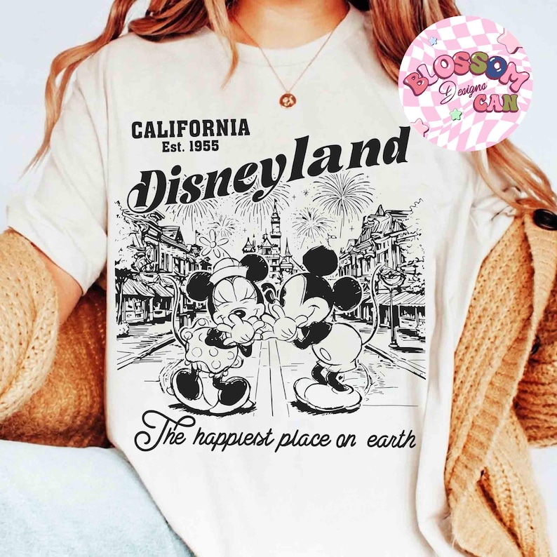 May include: Cream-colored t-shirt featuring a black and white graphic of Mickey and Minnie Mouse in front of a Disneyland scene. The shirt includes the text "CALIFORNIA Est. 1955 Disneyland" and "The happiest place on earth."