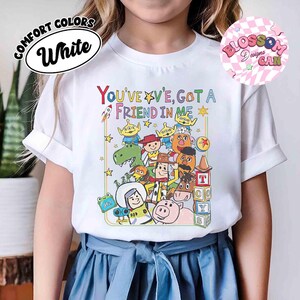 May include: White Comfort Colors t-shirt featuring a colorful graphic of characters and the text "YOU'VE GOT A FRIEND IN ME". The design includes a rocket ship, stars, and toy blocks. A Blossom Designs CAN logo is in the upper right.