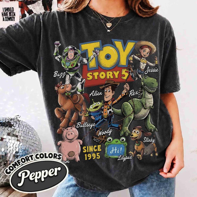 May include: A charcoal gray short-sleeve t-shirt featuring a graphic print of Toy Story characters including Buzz, Woody, Jessie, Bullseye, Rex, Slinky, and Hamm. The text reads Toy Story 5, Since 1995, and Hi! with character names labeled.