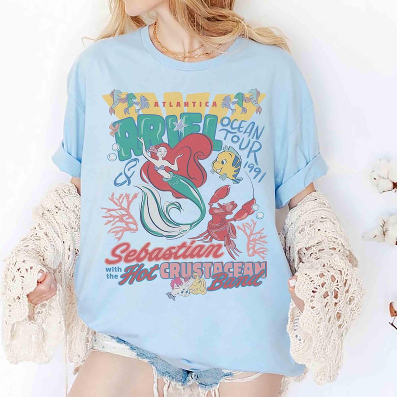 May include: Light blue t-shirt featuring a graphic of Ariel from The Little Mermaid, with the text "Ariel Ocean Tour 1991". The design includes illustrations of Sebastian, Flounder, and coral. The shirt has rolled-up sleeves.