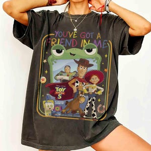 May include: A charcoal gray short-sleeve t-shirt featuring a graphic print with the text "YOU'VE GOT A FRIEND IN ME" and "TOY STORY 5 IN THEATERS JUNE 19". The design includes illustrations of Buzz Lightyear, Woody, Jessie, Bullseye, and a green frog frame.