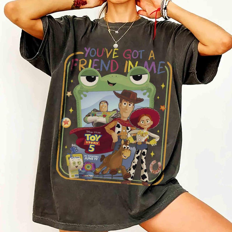 May include: A charcoal gray short-sleeve t-shirt featuring a graphic print with the text "YOU'VE GOT A FRIEND IN ME" and "TOY STORY 5 IN THEATERS JUNE 19". The design includes illustrations of Buzz Lightyear, Woody, Jessie, Bullseye, and a green frog frame.