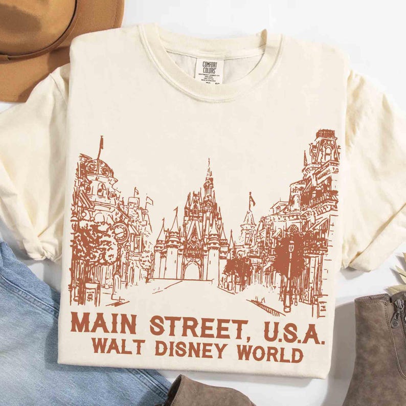 Peut inclure: T-shirt cr&egrave;me avec une illustration marron de Main Street, U.S.A., avec le ch&acirc;teau embl&eacute;matique au centre. Le texte "MAIN STREET, U.S.A. WALT DISNEY WORLD" est imprim&eacute; en dessous. Le t-shirt est en mati&egrave;re douce.