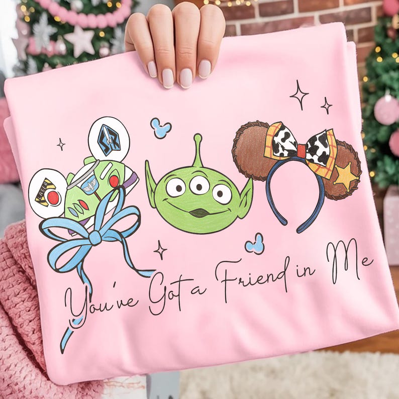 May include: Pink t-shirt featuring cartoon graphics of Buzz Lightyear, a Little Green Alien, and a headband with a cow print bow and star. The text "You've Got a Friend in Me" is written in script.