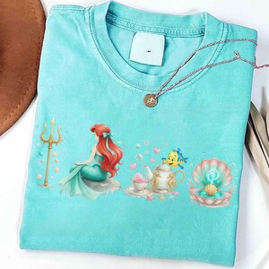 May include: A light blue t-shirt featuring a colorful illustration of a mermaid, a trident, and a seashell. The design includes a teapot, teacup, and a fish. A gold necklace with a pendant is on the shirt.