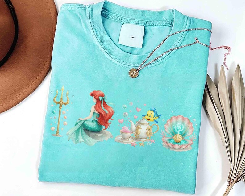May include: A light blue t-shirt featuring a colorful illustration of a mermaid, a trident, and a seashell. The design includes a teapot, teacup, and a fish. A gold necklace with a pendant is on the shirt.