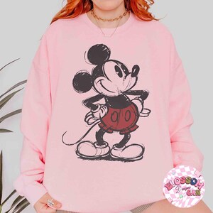 Op de afbeelding: Lichtroze sweatshirt met een zwart-rode Mickey Mouse afbeelding. Het stripfiguur lacht met zijn handen op zijn heupen. De sweatshirt heeft een ronde hals en lange mouwen. De afbeelding is in vintage stijl.