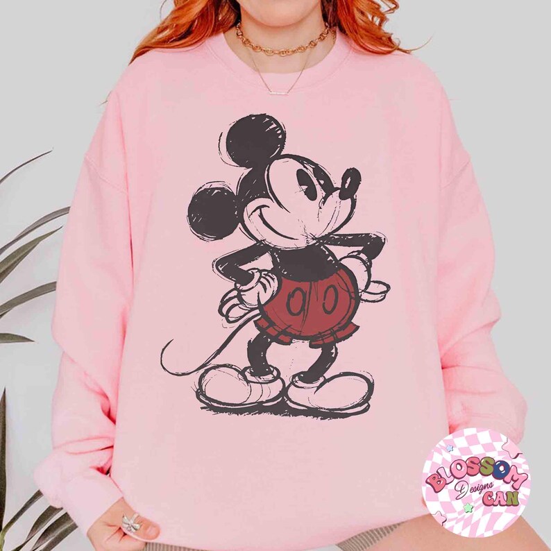 Op de afbeelding: Lichtroze sweatshirt met een zwart-rode Mickey Mouse afbeelding. Het stripfiguur lacht met zijn handen op zijn heupen. De sweatshirt heeft een ronde hals en lange mouwen. De afbeelding is in vintage stijl.