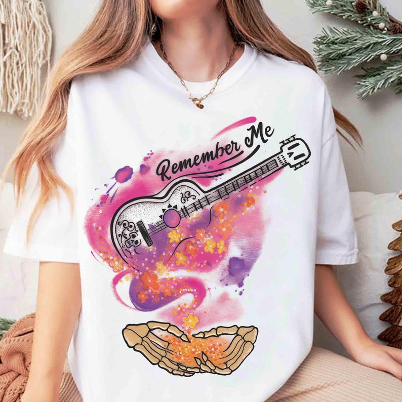 Puede incluir: Camiseta blanca con un dise&ntilde;o de guitarra y el texto "Remember Me". La guitarra est&aacute; decorada con motivos florales y de calaveras, sobre un fondo de acuarela en tonos rosa, morado y naranja. Una mano esquel&eacute;tica en la parte inferior.