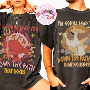 May include: Two dark gray t-shirts with cartoon graphics and text. One shirt features a red character with a trident and the text "I'M GONNA LEAD YOU DOWN THE PATH THAT ROCKS." The other shirt has a tan character and the text "I'M GONNA LEAD YOU DOWN THE PATH OF RIGHTEOUSNESS."