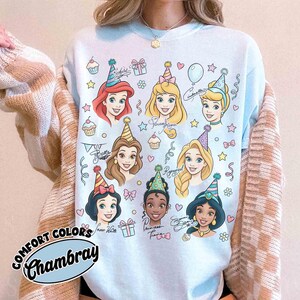 May include: Light blue sweatshirt featuring cartoon illustrations of Disney princesses wearing party hats. The design includes cupcakes, stars, and gift boxes. The text "COMFORT COLORS Chambray" is printed on the bottom left.