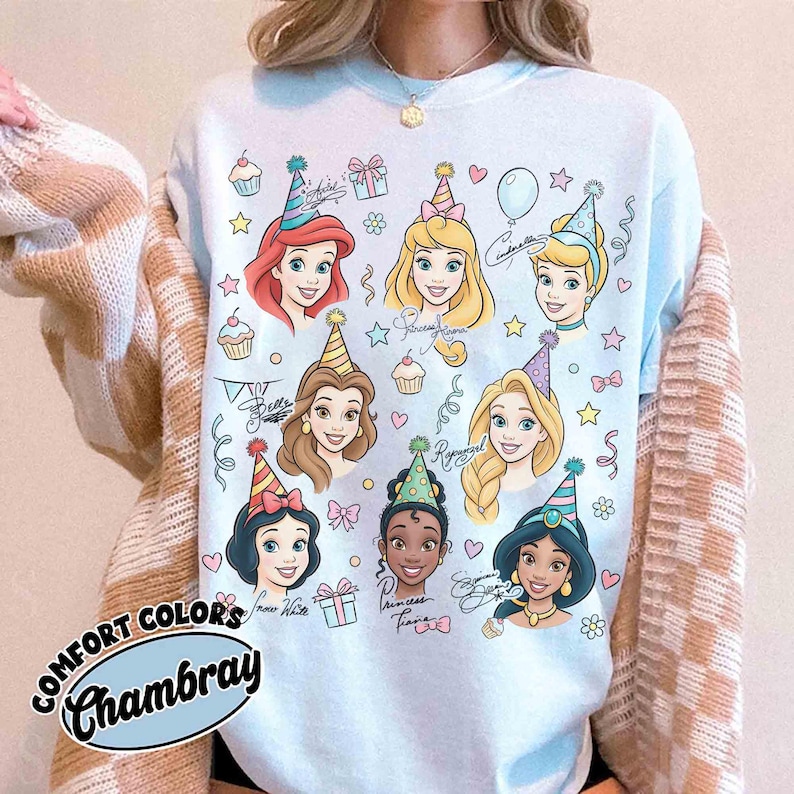 May include: Light blue sweatshirt featuring cartoon illustrations of Disney princesses wearing party hats. The design includes cupcakes, stars, and gift boxes. The text "COMFORT COLORS Chambray" is printed on the bottom left.