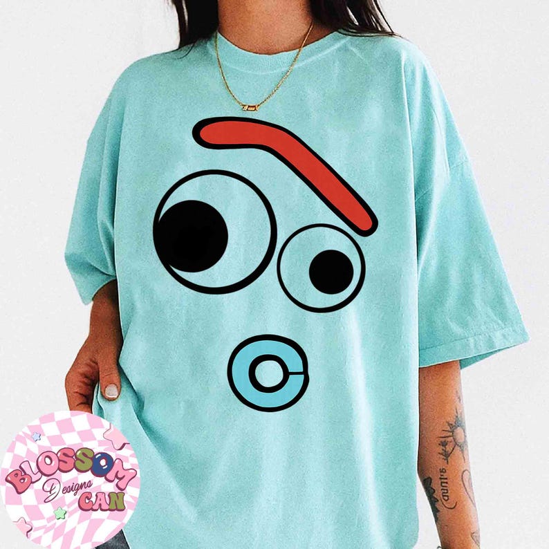 May include: Light blue t-shirt featuring a cartoon face design with large black and white eyes, a red eyebrow, and a blue circle with a white center. The shirt has short sleeves and a relaxed fit. The Blossom Designs CAN logo is visible.