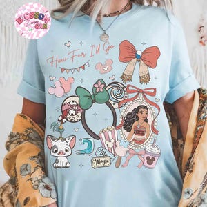 May include: Light blue t-shirt with the text "How Far I'll Go" and various cartoon illustrations. The design includes a portrait, bows, Mickey Mouse ears, and other themed graphics. The shirt has rolled-up sleeves.