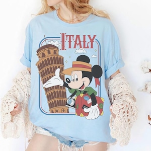 Pode incluir: Camiseta azul clara com um gr&aacute;fico do Mickey Mouse em frente &agrave; Torre de Pisa e a palavra "Italy". Mickey est&aacute; segurando uma c&acirc;mera e uma mala. O texto "Italy" &eacute; vermelho. A camiseta tem mangas enroladas.