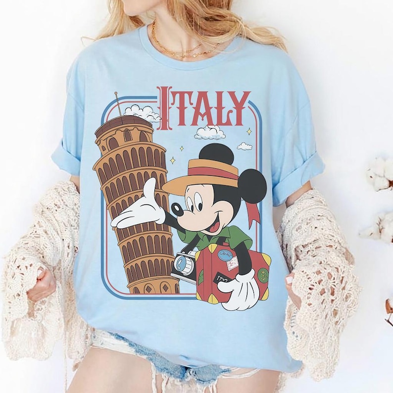Pode incluir: Camiseta azul clara com um gr&aacute;fico do Mickey Mouse em frente &agrave; Torre de Pisa e a palavra "Italy". Mickey est&aacute; segurando uma c&acirc;mera e uma mala. O texto "Italy" &eacute; vermelho. A camiseta tem mangas enroladas.