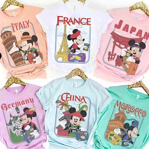 Pode incluir: Seis camisetas coloridas com designs tem&aacute;ticos de viagens com Mickey Mouse. Cada camisa tem o nome de um pa&iacute;s diferente: It&aacute;lia, Fran&ccedil;a, Jap&atilde;o, Alemanha, China e Marrocos. As camisas s&atilde;o exibidas em cabides dourados.