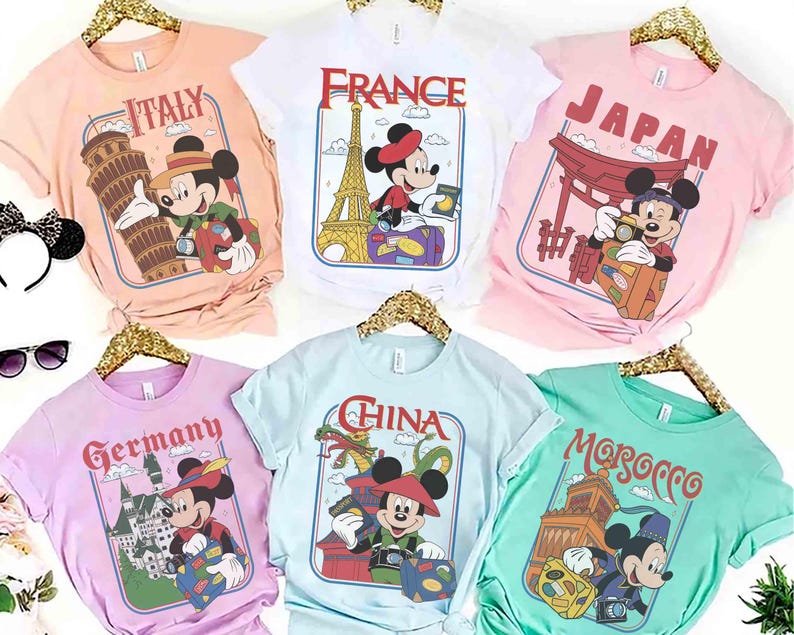 Pode incluir: Seis camisetas coloridas com designs tem&aacute;ticos de viagens com Mickey Mouse. Cada camisa tem o nome de um pa&iacute;s diferente: It&aacute;lia, Fran&ccedil;a, Jap&atilde;o, Alemanha, China e Marrocos. As camisas s&atilde;o exibidas em cabides dourados.