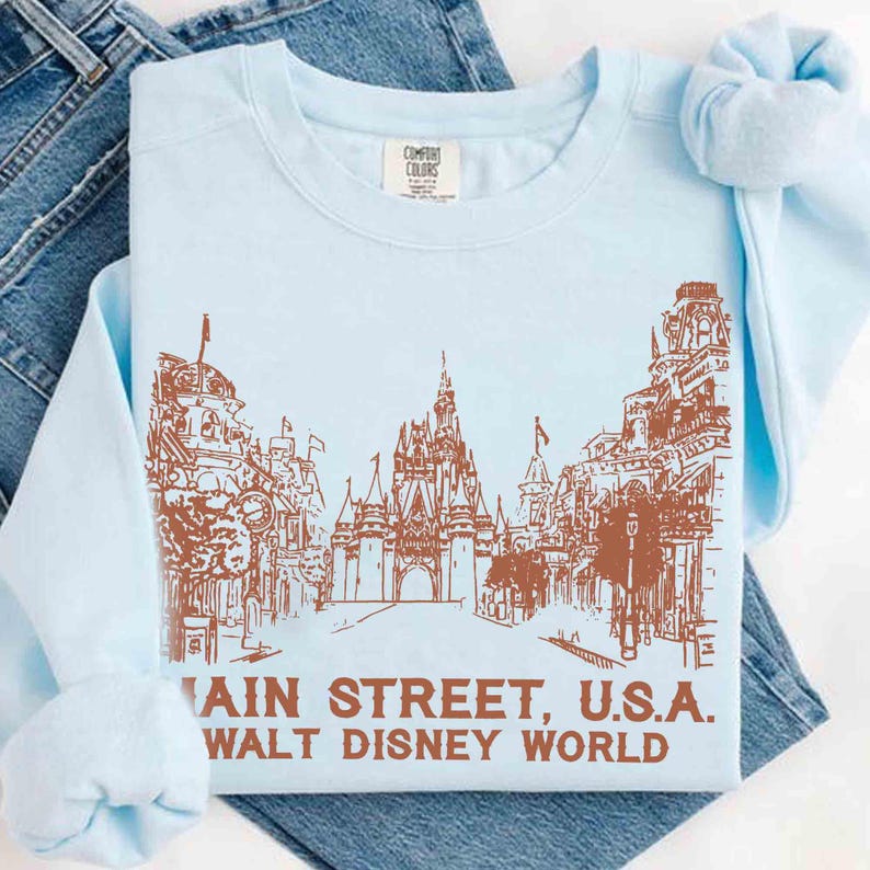Peut inclure: Un sweat-shirt bleu clair avec une illustration marron de Main Street, U.S.A. &agrave; Walt Disney World. Le motif comprend des b&acirc;timents, un ch&acirc;teau et le texte "MAIN STREET, U.S.A. WALT DISNEY WORLD" en marron.