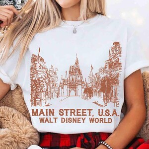 Peut inclure: T-shirt blanc avec un dessin marron de Main Street, U.S.A. et du ch&acirc;teau de Cendrillon de Walt Disney World. Le texte "MAIN STREET, U.S.A. WALT DISNEY WORLD" est imprim&eacute; en dessous. Le t-shirt a des manches courtes.