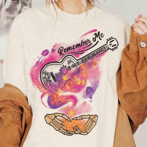 Puede incluir: Camiseta color crema con un gr&aacute;fico colorido de una guitarra y el texto "Remember Me". El dise&ntilde;o de la guitarra incorpora elementos florales y de calaveras, sobre un vibrante fondo de acuarela rosa y morado. Un par de manos esquel&eacute;ticas en la parte inferior.