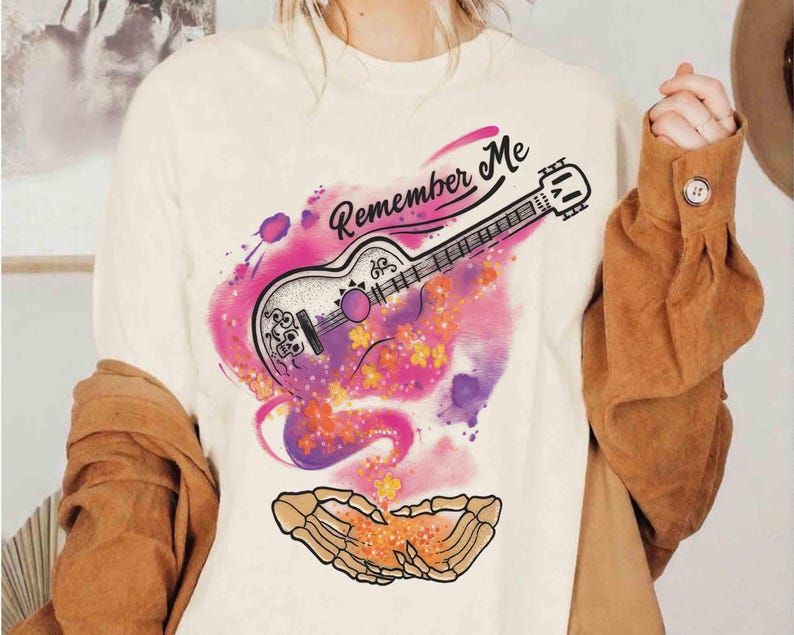 Puede incluir: Camiseta color crema con un gr&aacute;fico colorido de una guitarra y el texto "Remember Me". El dise&ntilde;o de la guitarra incorpora elementos florales y de calaveras, sobre un vibrante fondo de acuarela rosa y morado. Un par de manos esquel&eacute;ticas en la parte inferior.