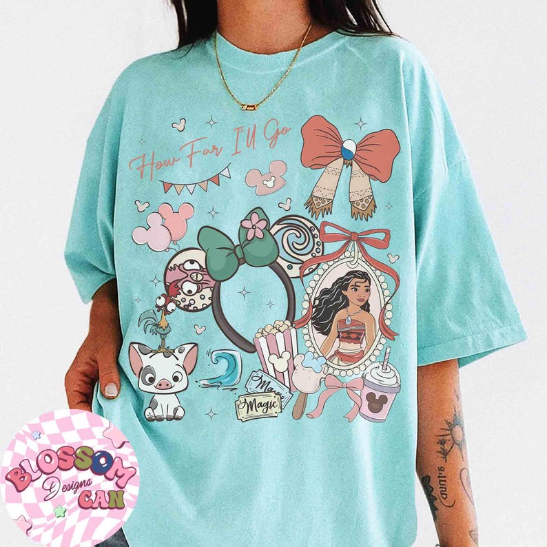 May include: A light blue t-shirt with a colorful graphic design featuring the text "How Far I'll Go" and various cartoon elements. The design includes a portrait, bows, and other whimsical illustrations.