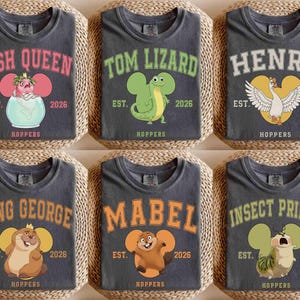 Puede incluir: Seis camisetas de color gris oscuro con varios diseños de personajes de dibujos animados. Cada camiseta presenta un nombre diferente en una fuente universitaria, junto con el texto "EST. 2026 HOPPERS". Los diseños incluyen un pez, un lagarto, un ganso, un castor y un insecto.
