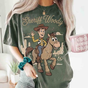 Op de afbeelding: Een donker olijfgroen t-shirt met een grafische print van Sheriff Woody op het paard Bullseye. De tekst Sheriff Woody staat in een cursief lettertype boven de figuren. Het ontwerp bevat een woestijnlandschap met cactussen.