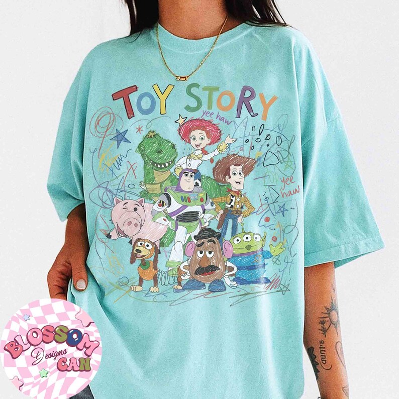 Puede incluir: Camiseta azul claro con la frase "Toy Story" y personajes de dibujos animados como Woody y Buzz Lightyear. El dise&ntilde;o tiene una est&eacute;tica dibujada a mano con acentos garabateados.