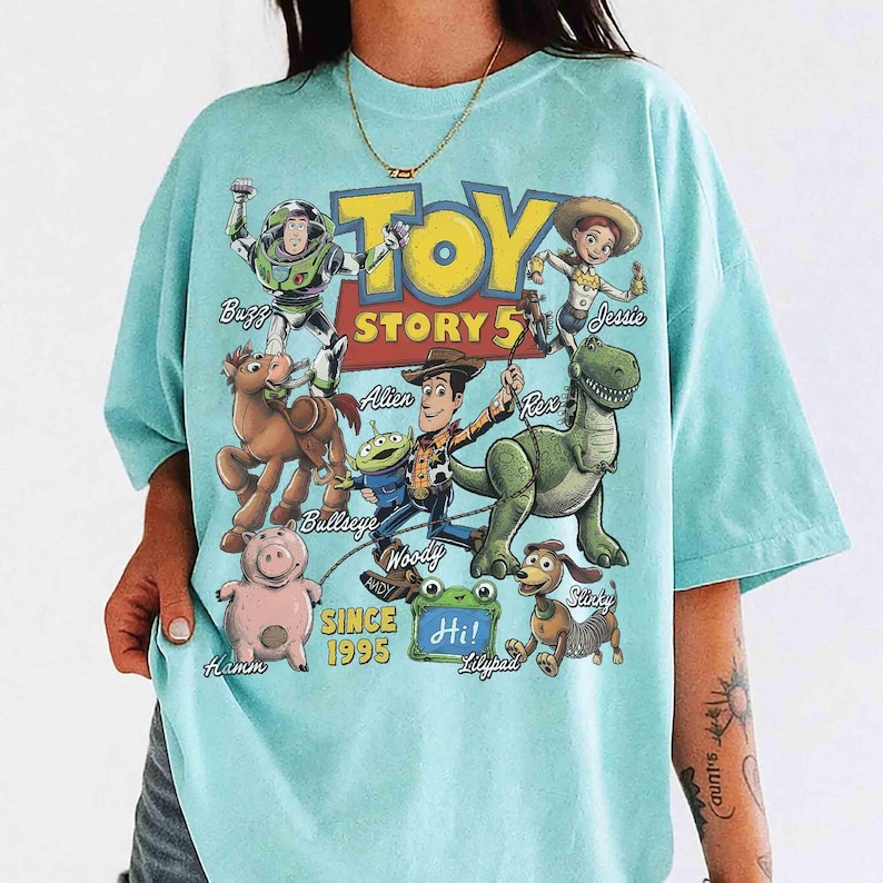 May include: A light blue t-shirt featuring a graphic print of Toy Story characters including Buzz Lightyear, Jessie, Bullseye, Woody, Rex, Hamm, Slinky, and an alien. The text reads Toy Story 5, Since 1995, and Hi! Lilypad.