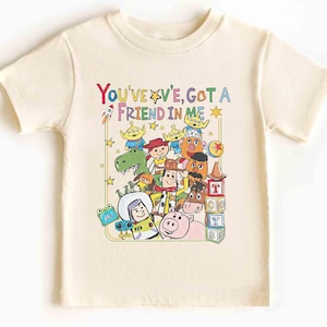 May include: Cream-colored t-shirt featuring a colorful graphic of characters and the text "You've Got A Friend In Me." The design includes a rocket, stars, and various cartoon figures. The shirt is short-sleeved and appears to be made of cotton.