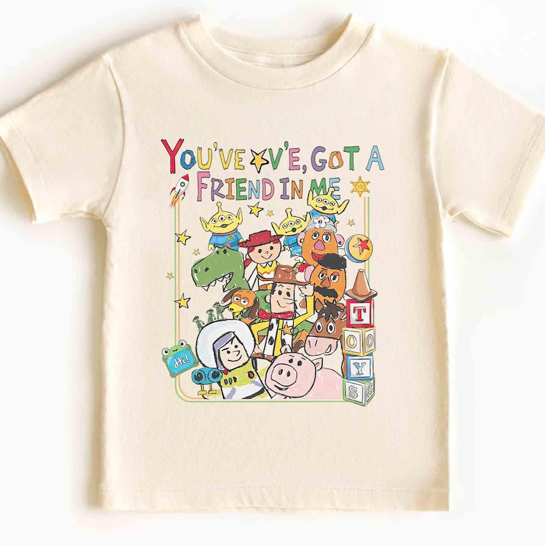May include: Cream-colored t-shirt featuring a colorful graphic of characters and the text "You've Got A Friend In Me." The design includes a rocket, stars, and various cartoon figures. The shirt is short-sleeved and appears to be made of cotton.