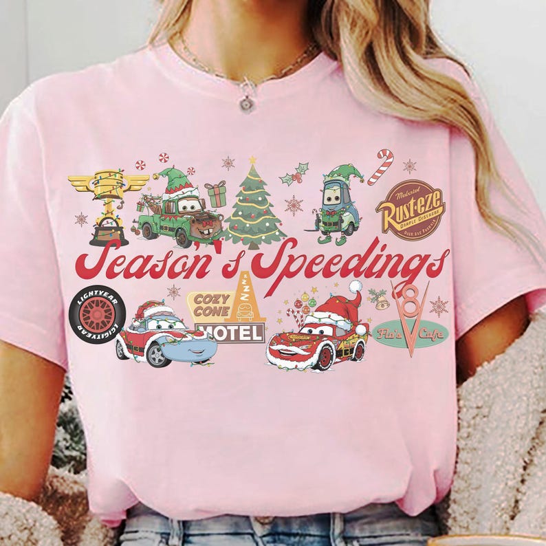 May include: Pink t-shirt featuring a festive design with characters from the movie Cars, including Lightning McQueen and Mater, in holiday attire. The text "Season's Speedings" is displayed in a festive font, with other holiday-themed graphics.