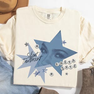 May include: Cream-colored t-shirt featuring a blue starburst design with the text "Silent Treatment" and the name "FREYA SKYE". The design includes an image of eyes within the starburst and small star accents.