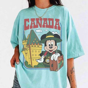 Pode incluir: Uma camiseta azul clara com a palavra "CANADA" em letras vermelhas. A camiseta apresenta uma imagem de desenho animado do Mickey Mouse vestido como guarda florestal, segurando uma c&acirc;mera e uma mala, com um castelo canadense ao fundo.