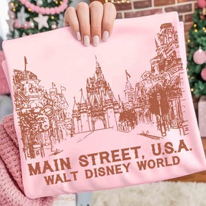 Peut inclure: Un t-shirt rose clair avec un dessin marron de Main Street, U.S.A. et du ch&acirc;teau de Cendrillon de Walt Disney World. Le texte "MAIN STREET, U.S.A. WALT DISNEY WORLD" est imprim&eacute; en marron en dessous.