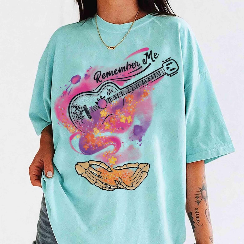 Puede incluir: Una camiseta azul claro con un gr&aacute;fico colorido. El dise&ntilde;o incluye una guitarra con el texto "Remember Me" encima, rodeada de salpicaduras rosas y moradas estilo acuarela. Debajo de la guitarra hay manos esquel&eacute;ticas sosteniendo flores.
