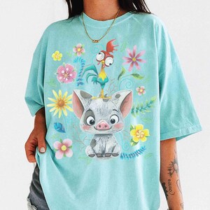 May include: A light blue t-shirt with a cartoon pig and rooster design, surrounded by flowers. The pig is grey and white, with a smiling expression. The rooster is red, yellow, and blue. The flowers are pink, yellow, and white.
