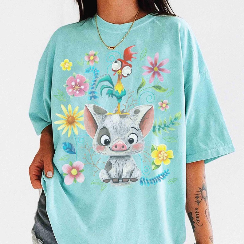 May include: A light blue t-shirt with a cartoon pig and rooster design, surrounded by flowers. The pig is grey and white, with a smiling expression. The rooster is red, yellow, and blue. The flowers are pink, yellow, and white.