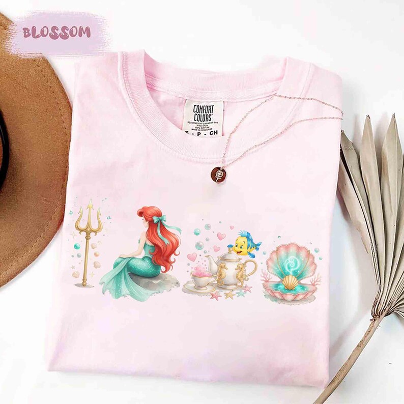 May include: Pink Comfort Colors t-shirt featuring a watercolor design of a mermaid, a teapot, and a seashell. The design includes a golden trident, a blue fish, and a necklace. The word "BLOSSOM" is printed in the upper left corner.