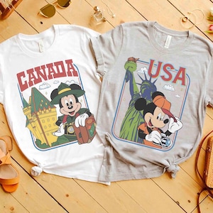 Pode incluir: Duas camisetas com gr&aacute;ficos tem&aacute;ticos de viagem. Uma &eacute; branca com "CANADA" e um rato de desenho animado com um chap&eacute;u verde. A outra &eacute; cinza com "USA" e um rato de desenho animado com um bon&eacute; laranja, com a Est&aacute;tua da Liberdade.